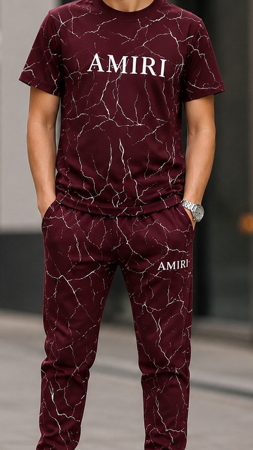 A man wearing a matching burgundy and white marble-patterned tracksuit with 'AMIRI' printed on the chest and thigh, paired with white sneakers, standing on a city sidewalk.