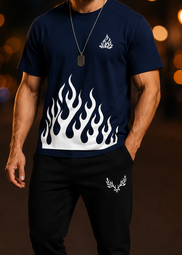 A man wearing a navy blue t-shirt with a white flame graphic and a small white logo on the chest, paired with black sweatpants, standing against a blurred night street background with warm bokeh lights.